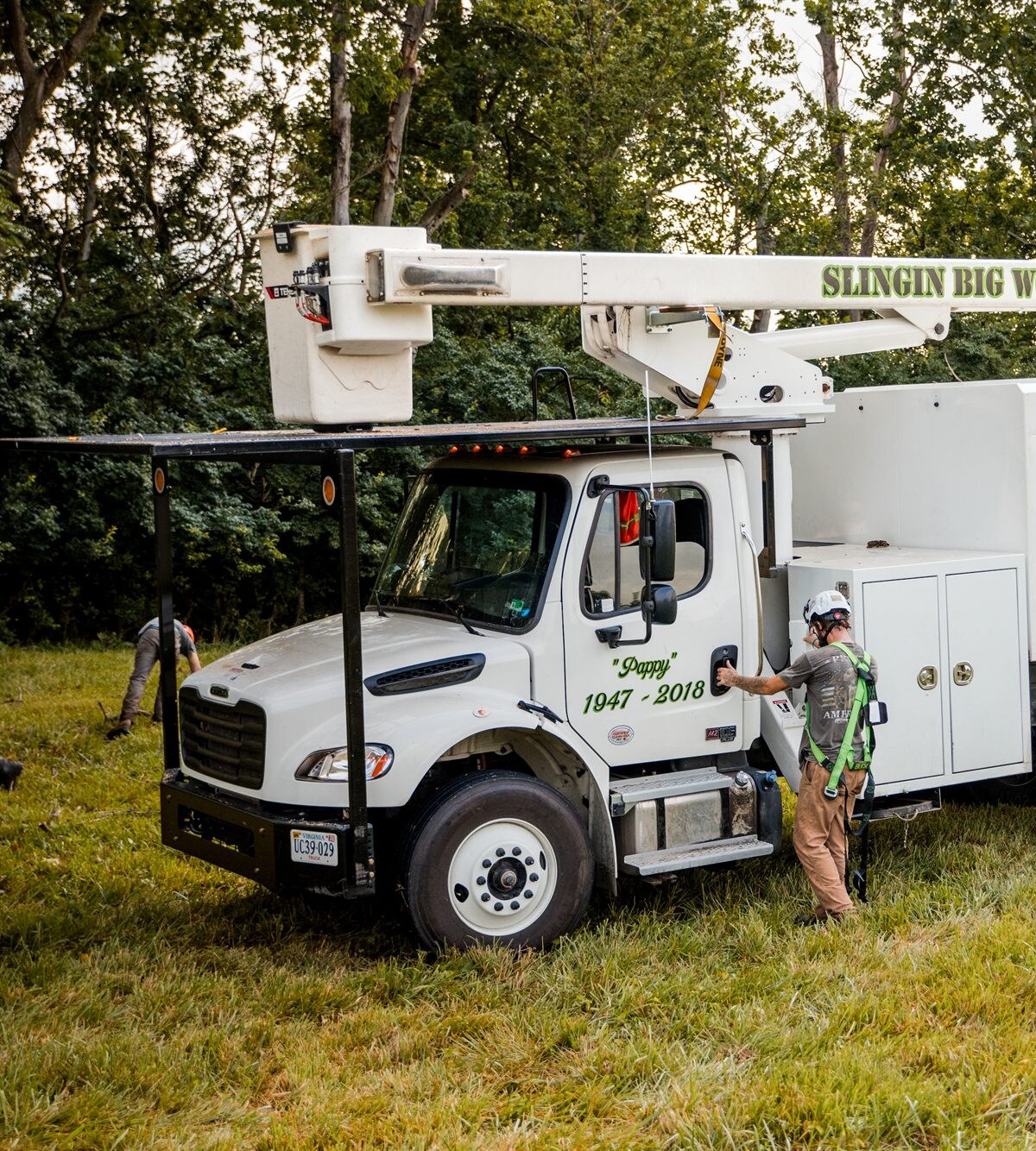 A white bucket truck parked on cut grass in front of a wood line with its boom stowed along the roof, a crew member in a green climbing harness and white helmet standing at an open compartment on the side of the truck, and a second worker crouching in the grass beyond the front bumper.