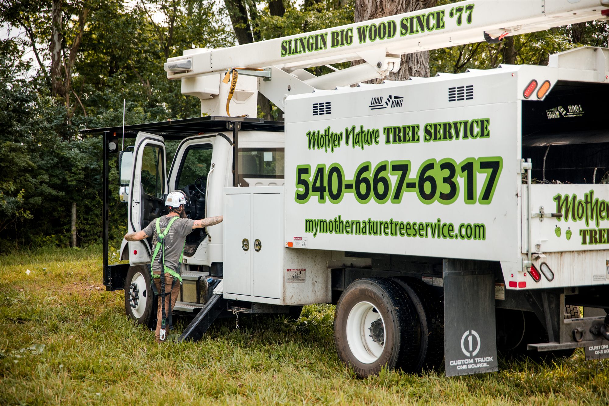 An arborist in a climbing harness standing at the open cab door of the bucket truck on grass